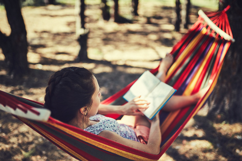 Female enjoying nature in the mountains, swinging in hammock, reading a book in the midst of trees. The mesmerizing tree shadows can be seen on the ground. Female swinging in hammock at a Tennessee Camping Destination; family friendly camping, cades cove camping