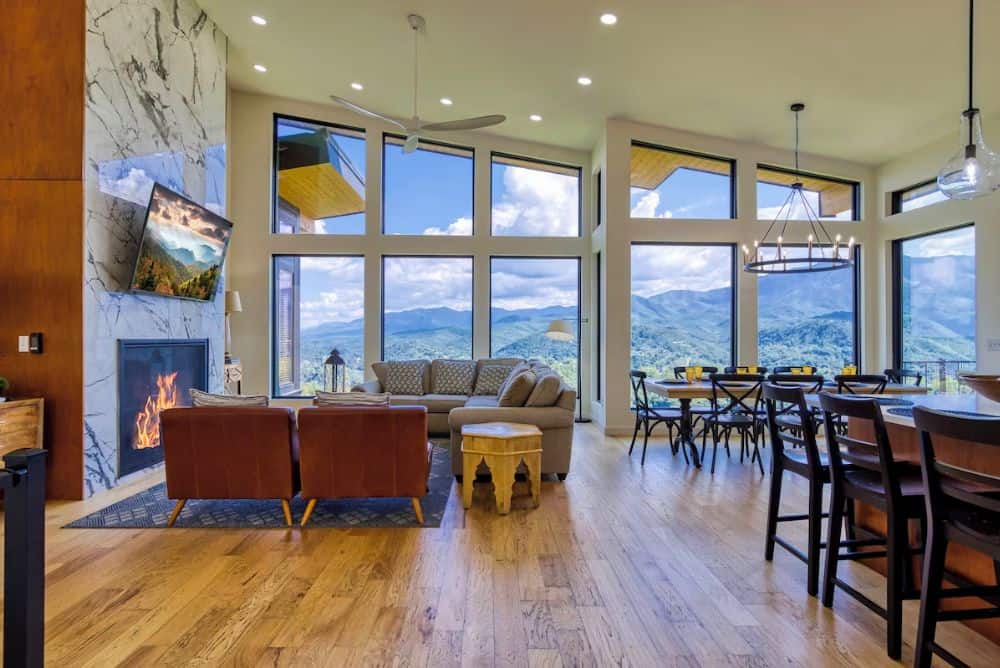 Spacious cabin living and dining area with floor-to-ceiling windows, fireplace, and panoramic Smoky Mountain views.
