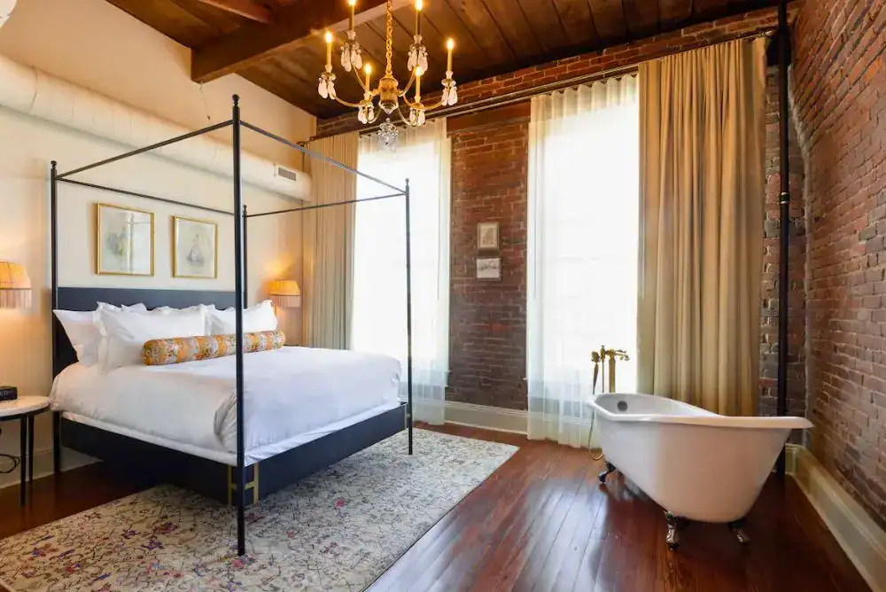 A boutique hotel room at Hotel Cleo in Knoxville featuring a black four-poster bed, exposed brick walls, hardwood floors, a clawfoot tub, and a chandelier overhead, a great experience for history lovers