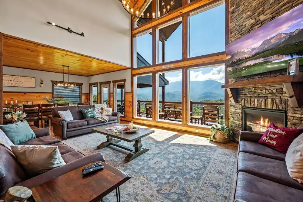 Great room at Mountain View Manor in Chalet Village with floor-to-ceiling windows, a stone fireplace, and panoramic Smoky Mountain views.