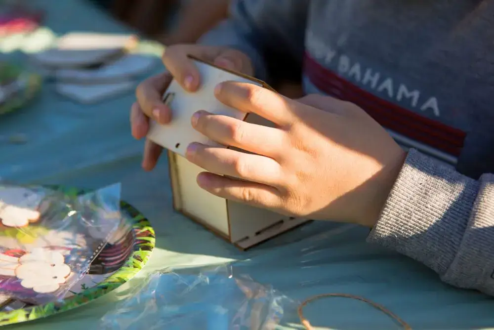 A child's hands piece together a wooden DIY birdhouse kit on an outdoor table.