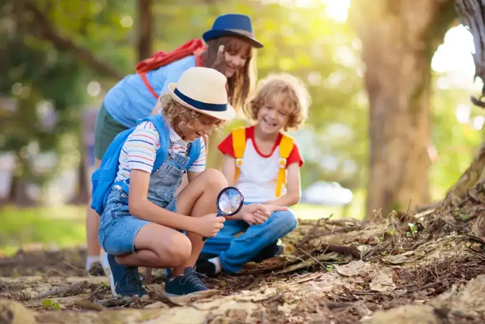 Three kids with backpacks crouch on a forest trail while one uses a magnifying glass to look closely at the ground.