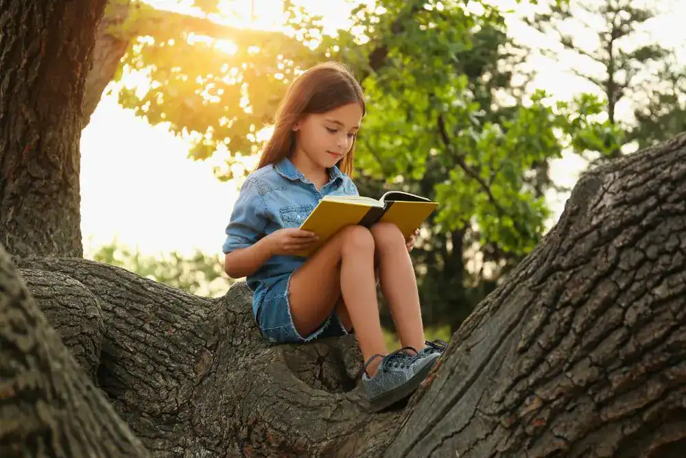 A young girl reads on a large tree branch in the evening sun, a reminder that books make great gifts for kids who love the outdoors.