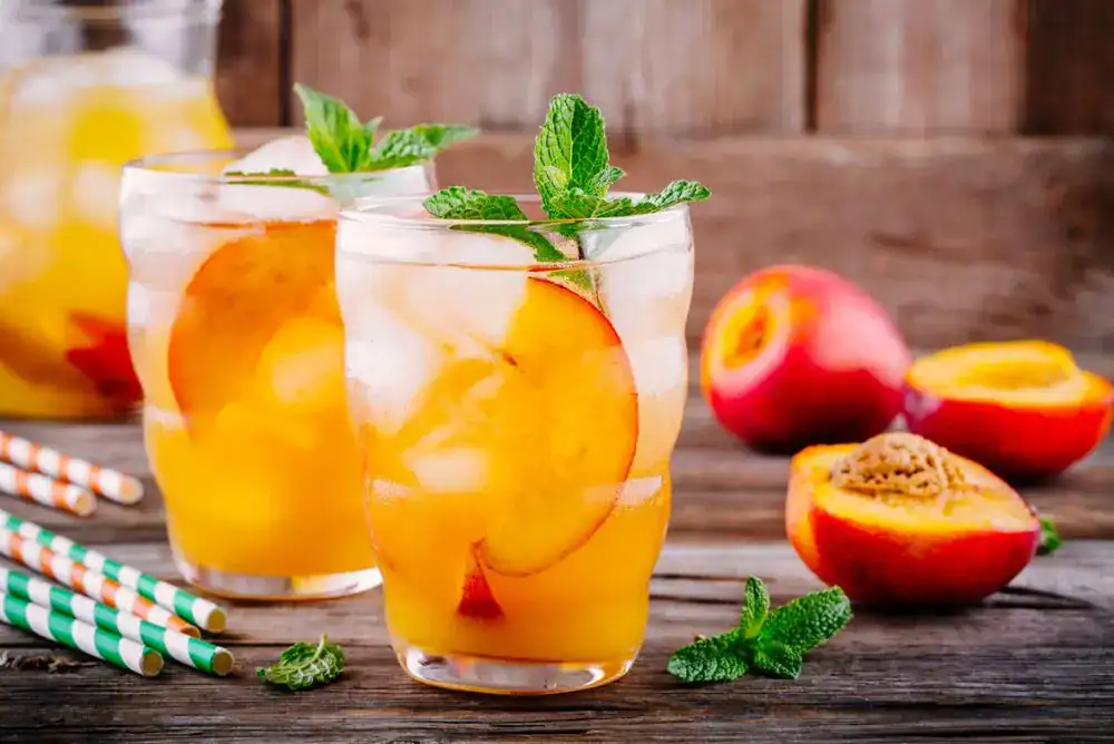 A glass of BLESS YOUR PEACH, a peach moonshine cocktail served over ice with a peach slice garnish—perfect for summer sipping.

