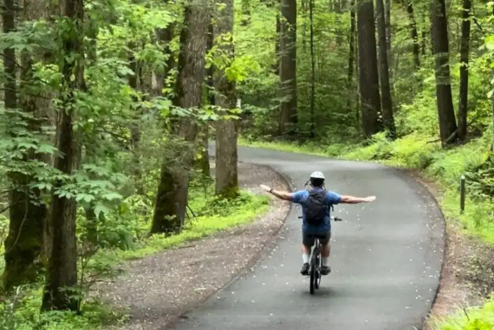Terry riding a bicycle on the scenic paved loop road in Cades Cove with mountains and trees in the background. One of our favorite things to do in the Smoky Mountains.