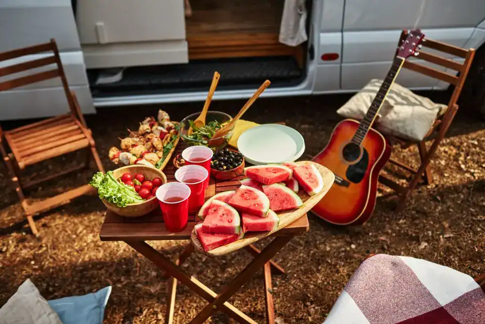 Summer camping picnic table with watermelon slices and campsite snacks outside an RV, a guitar nearby, and outdoor chairs at a campground.