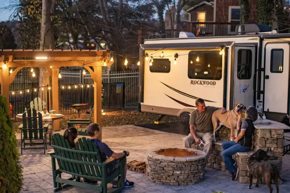 Family relaxing by a fire pit outside an RV at a KOA campground with outdoor seating and string lights in East Tennessee.