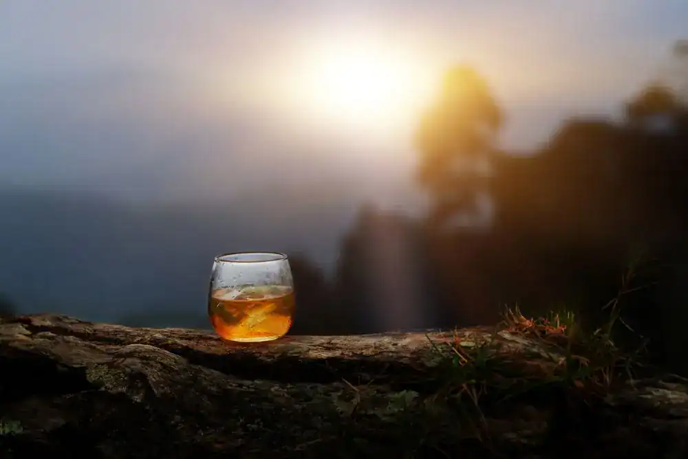 A glass of Tennessee whiskey resting on a rock ledge at sunset, overlooking a scenic outdoor Tennessee landscape

