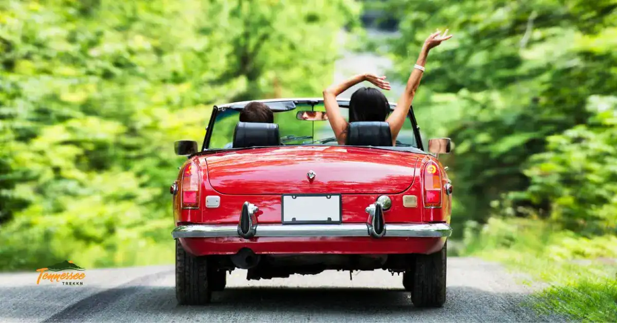 A couple riding in a red convertible down a tree-lined road, hands raised in the air on a Tennessee weekend getaway