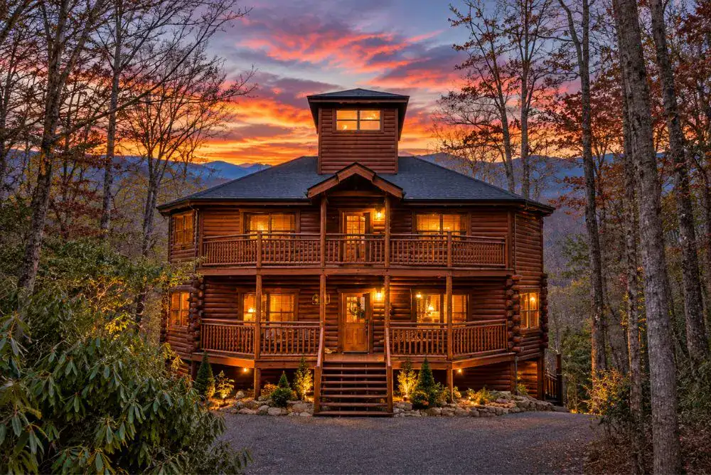 Big Dipper Lodge log cabin in Cosby, Tennessee, featuring two levels of wraparound porches, warm exterior lighting, and a colorful sunset.