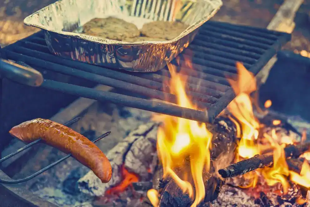 Sausage cooking on a campfire grill grate with a foil pan of burgers, a reminder to clean cooking gear for bear safety while camping