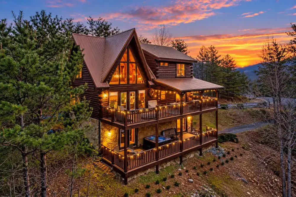 Large log cabin in Wears Valley near the Smoky Mountains, with wraparound decks, warm exterior lighting, and a colorful sunset.
