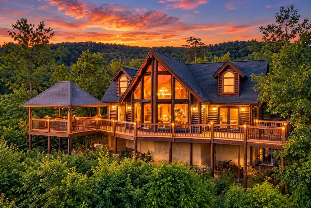 Summit's Peak log cabin in Wears Valley, Tennessee, featuring large windows, wraparound decks, and warm evening lights against a colorful sunset.